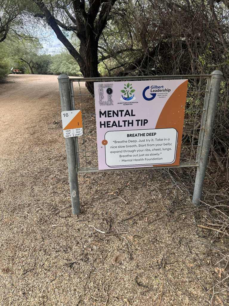 A sign offering a mental health tip about deep breathing at the Riparian Preserve at Water Ranch in Gilbert, Arizona, surrounded by trees and a gravel path.